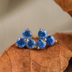 14k yellow gold stud earrings with 3 blue sapphires arranged in a floral shape, sitting on a brown leaf