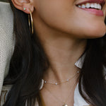 A close-up lifestyle shot of a woman wearing layered gold necklaces, including a delicate chain with tiny pearl-like beads and a leaf-shaped pendant, along with a gold hoop earring.