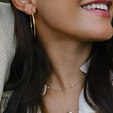 A close-up lifestyle shot of a woman wearing layered gold necklaces, including a delicate chain with tiny pearl-like beads and a leaf-shaped pendant, along with a gold hoop earring.