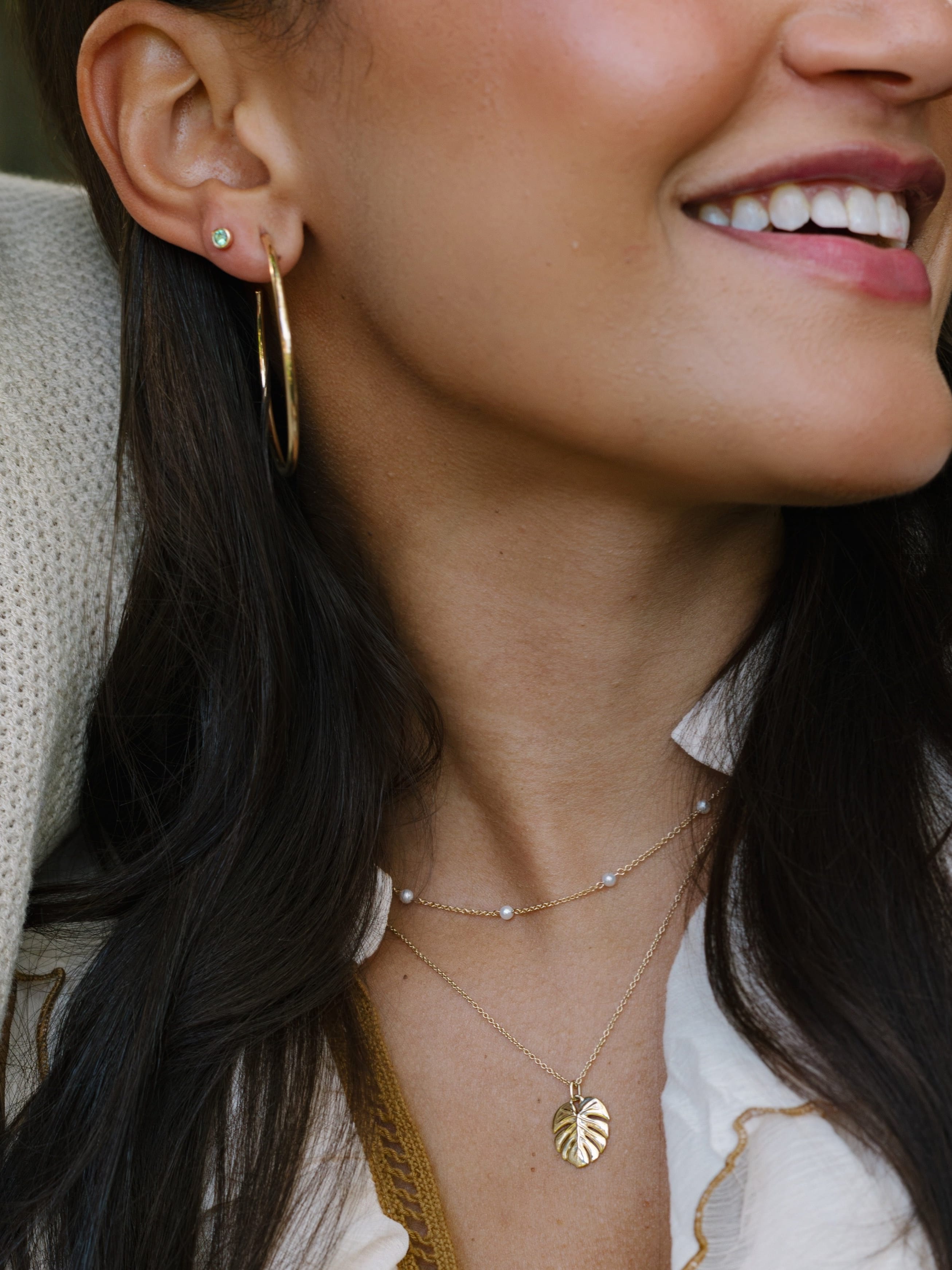 A close-up lifestyle shot of a woman wearing layered gold necklaces, including a delicate chain with tiny pearl-like beads and a leaf-shaped pendant, along with a gold hoop earring.