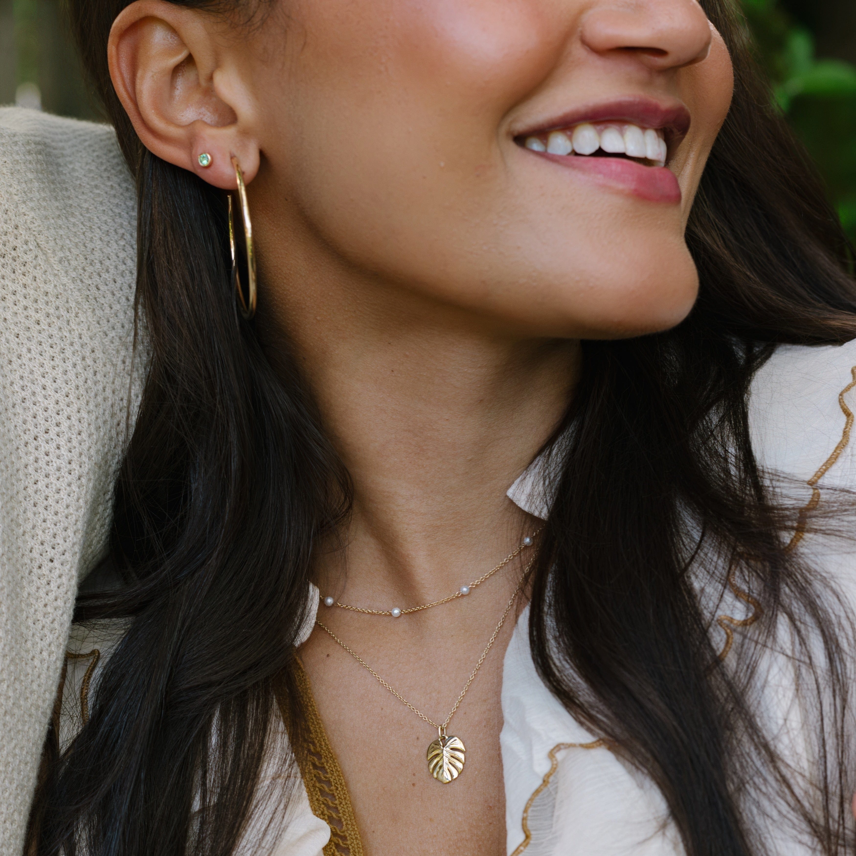 A close-up lifestyle shot of a woman wearing layered gold necklaces, including a delicate chain with tiny pearl-like beads and a leaf-shaped pendant, along with a gold hoop earring.