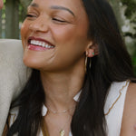 A close-up lifestyle shot of a smiling woman with long dark hair laughing, wearing gold hoop earrings, layered gold necklaces, and a cream sleeveless top, next to a man in a beige knit shirt.