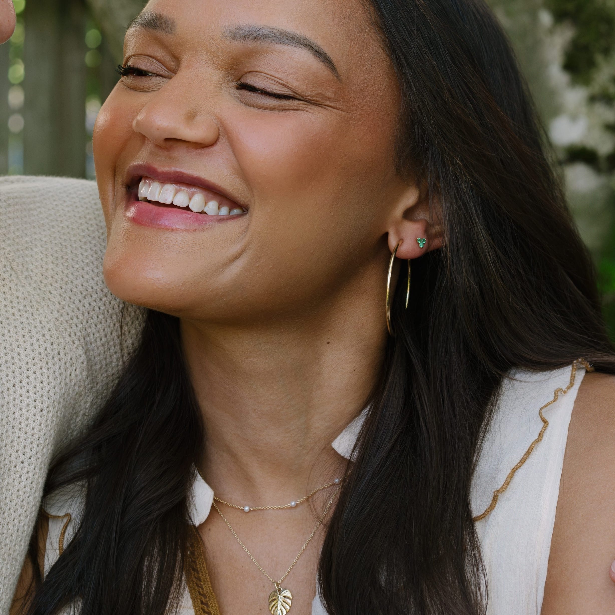 A close-up lifestyle shot of a smiling woman with long dark hair laughing, wearing gold hoop earrings, layered gold necklaces, and a cream sleeveless top, next to a man in a beige knit shirt.